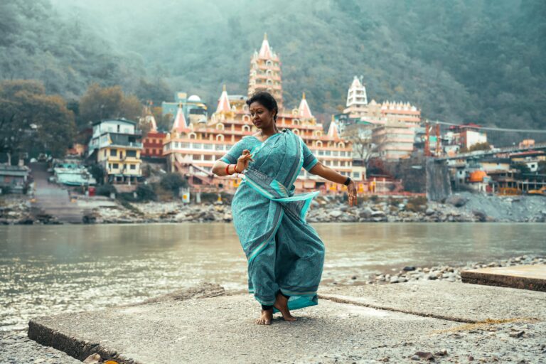 Graceful woman in traditional attire dancing by river in Rishikesh with scenic temples in background.