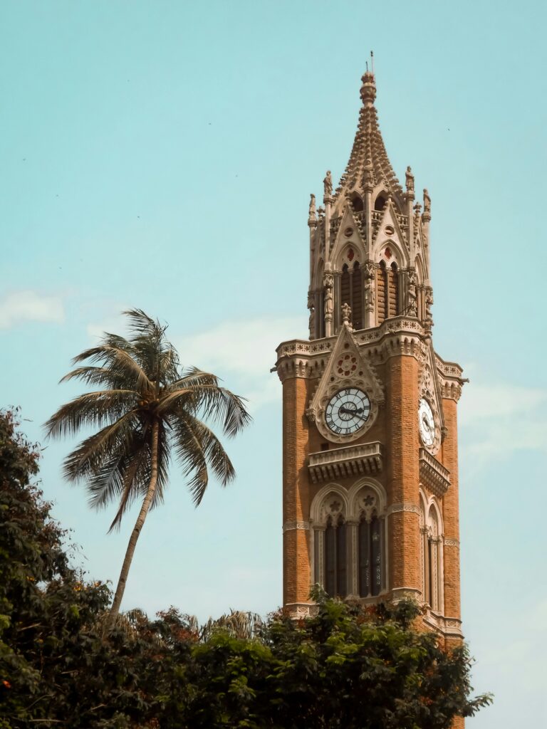 Gothic-style Rajabai Clock Tower stands tall amidst lush greenery and clear skies in Mumbai.
