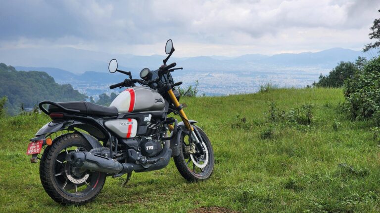 A motorcycle parked on a grassy hilltop overlooking a cityscape with distant mountains.
